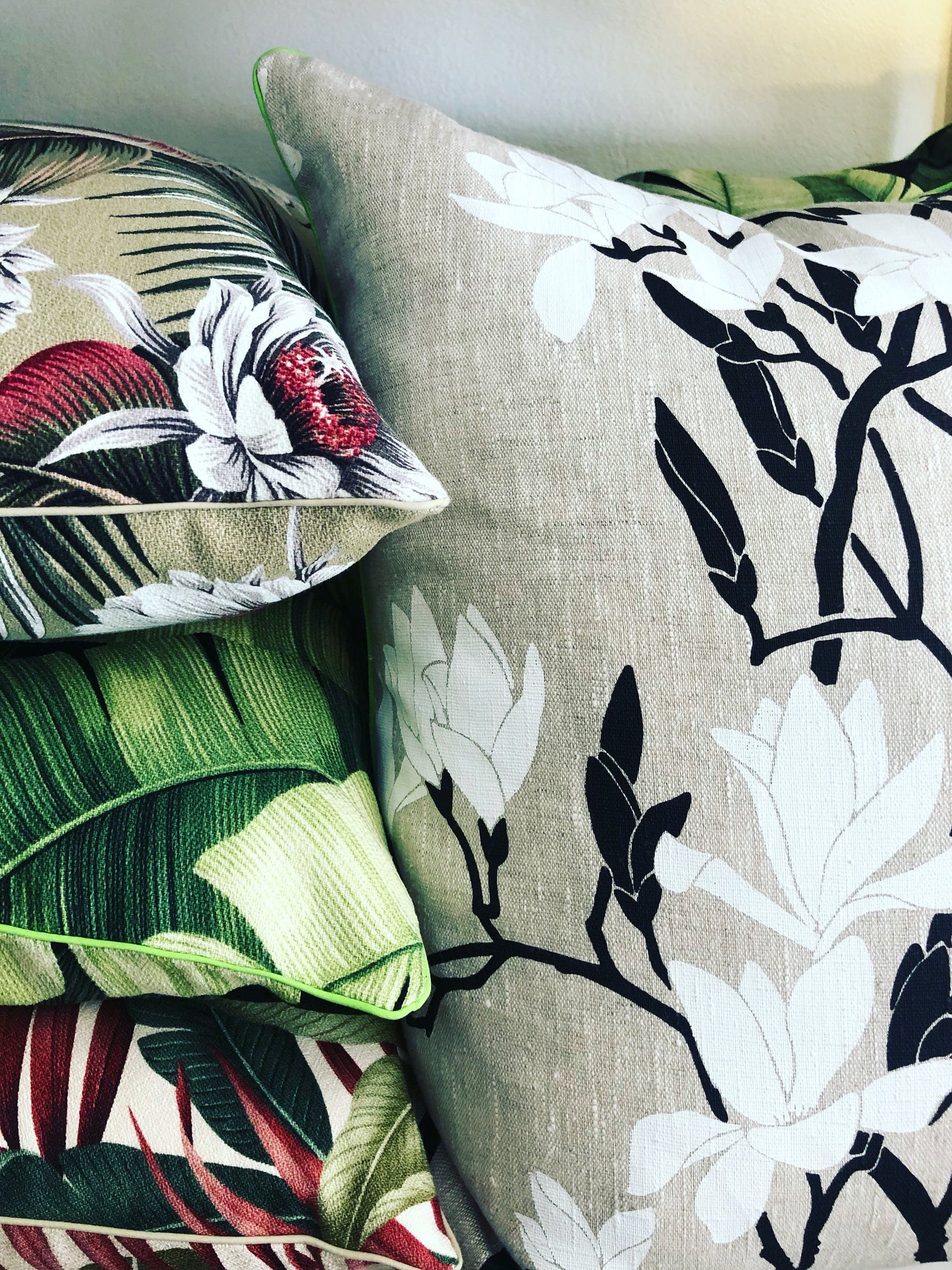 A set of four cushions with floral patterns, featuring magnolia flowers, in various shades of green, beige, and black, stacked on a sofa.