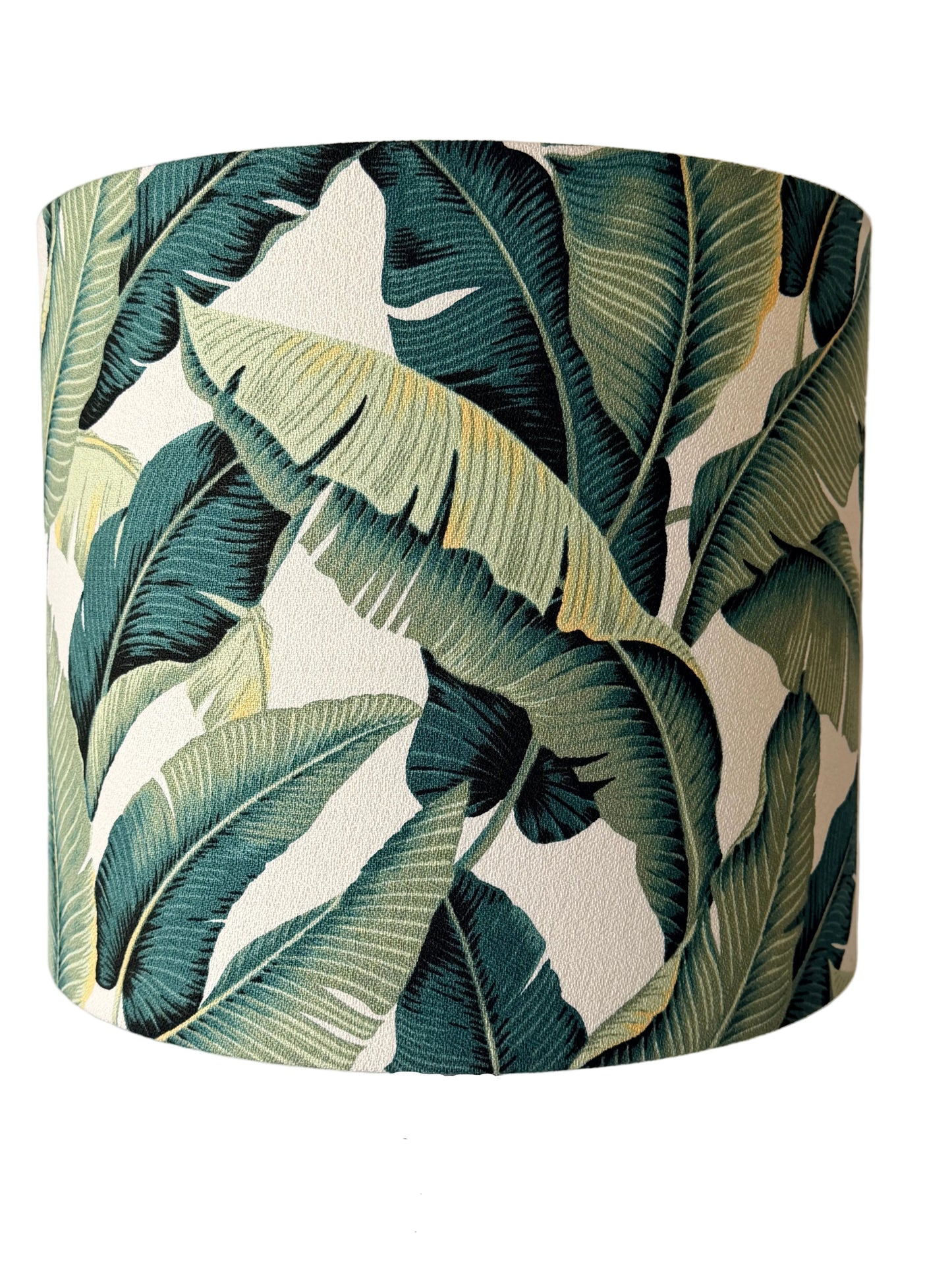 A cylindrical table lamp shade with a tropical banana leaf pattern in shades of green and beige,