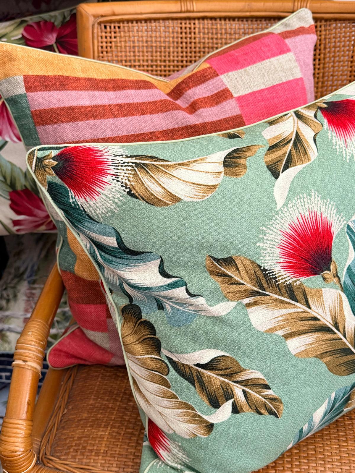 A square decorative throw pillow with a tropical floral and leaf print in sage green, red, brown, and cream, styled on a woven rattan chair next to another striped pillow.