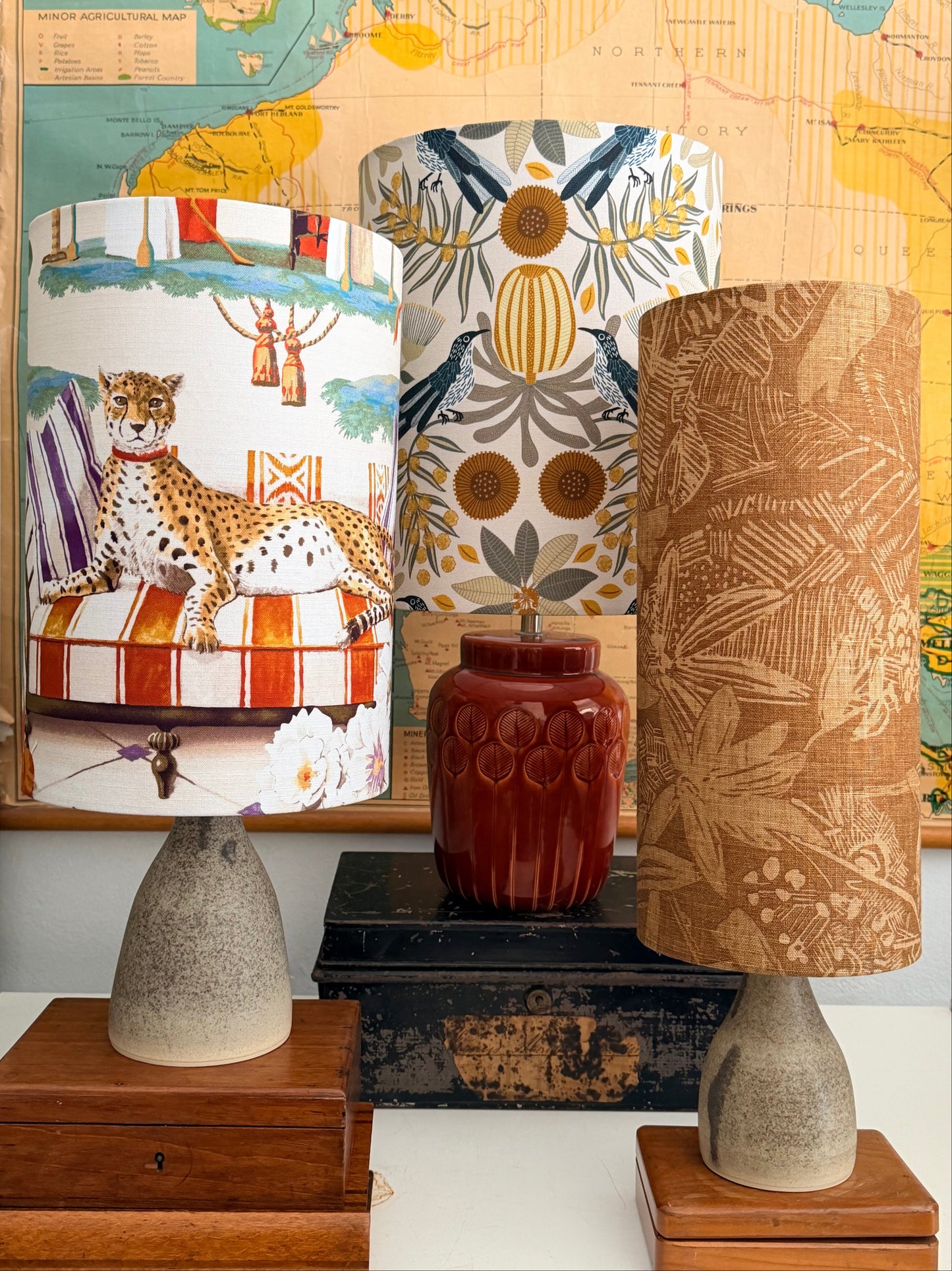 Three cylindrical lampshades with distinct patterns (brown palm leaves, blue and beige tropical leaves, and a leopard print with stripes) displayed next to a dark red ceramic vase and a vintage map.