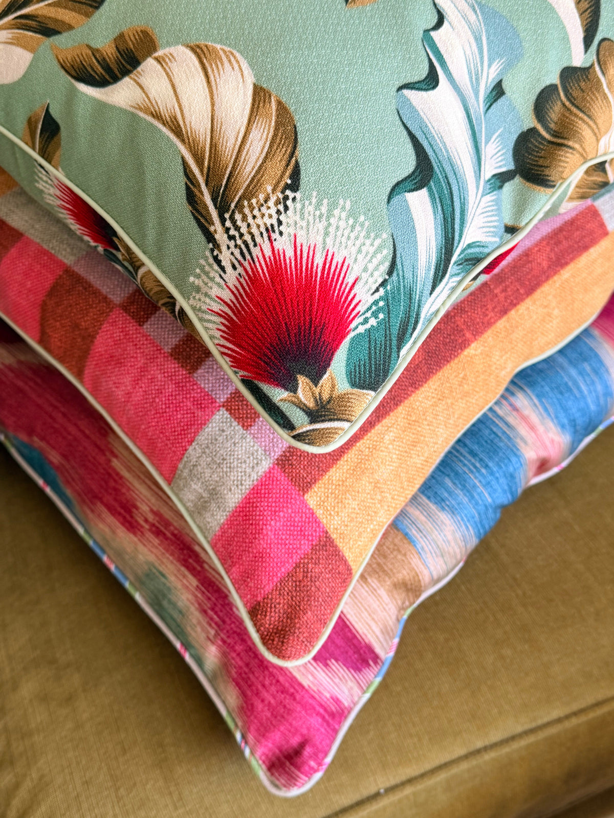 Two decorative throw pillows stacked on a tan upholstered chair. The top pillow features a sage green background with a large floral and leaf pattern in brown, teal, and red.