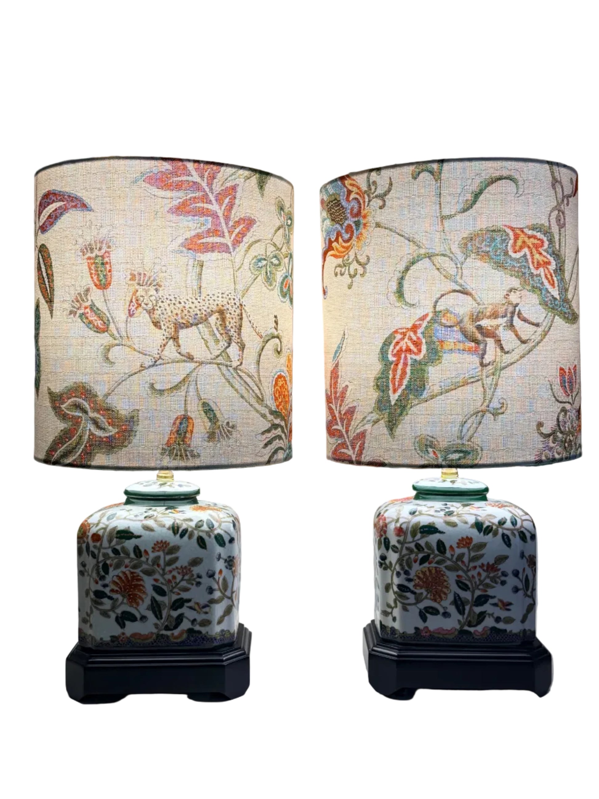 A pair of vintage-style table lamps with white fabric drum shades featuring a colorful botanical and animal print, and matching white ceramic jar bases with floral patterns