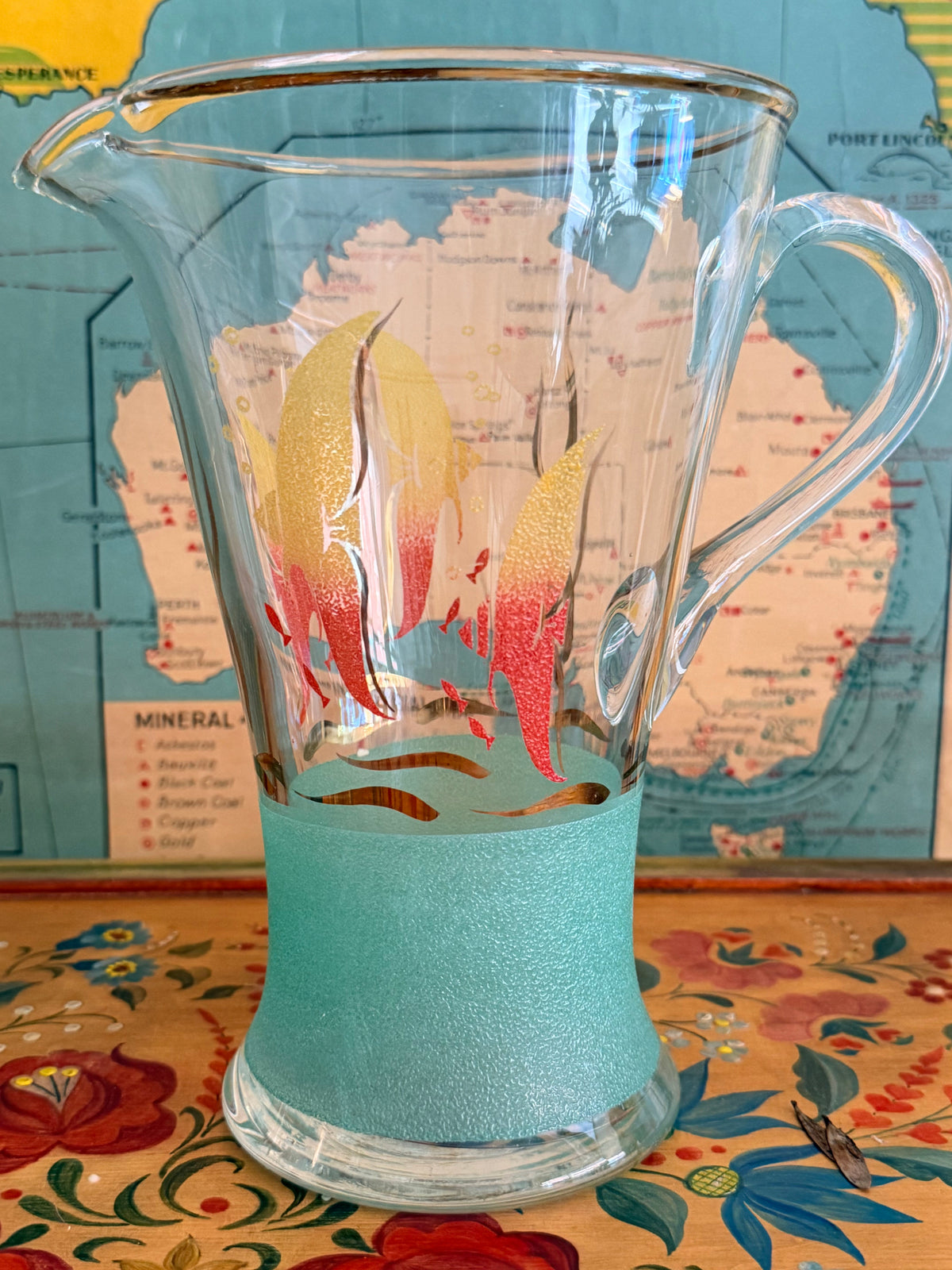 A  retro frosted pitcher with fotumblers, decorated with colorful fish illustrations in yellow, orange, and red and with a sea green frosted band