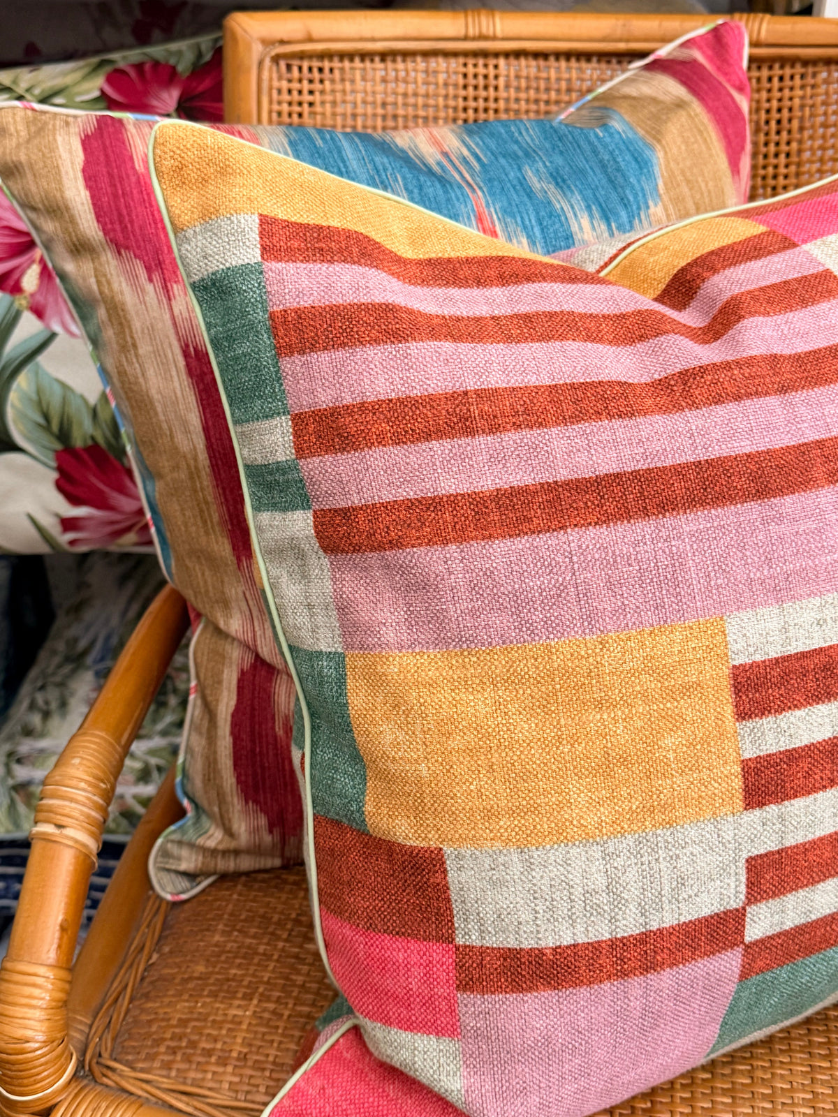 A close-up of a square throw pillow with a colorful patchwork block pattern in pink, orange, yellow, green, and beige, resting on a woven rattan chair.