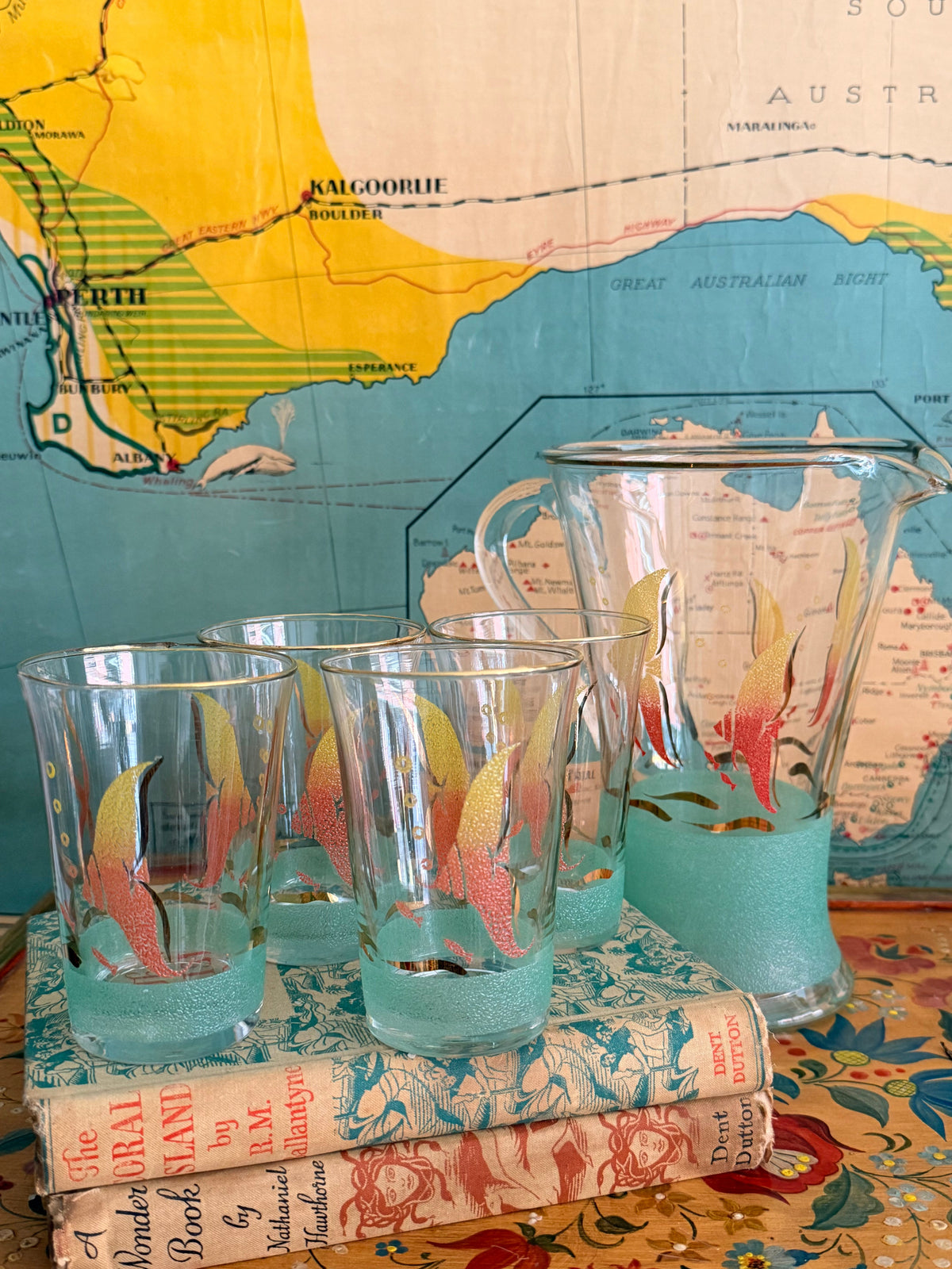 A  retro frosted pitcher with fotumblers, decorated with colorful fish illustrations in yellow, orange, and red and with a sea green frosted band