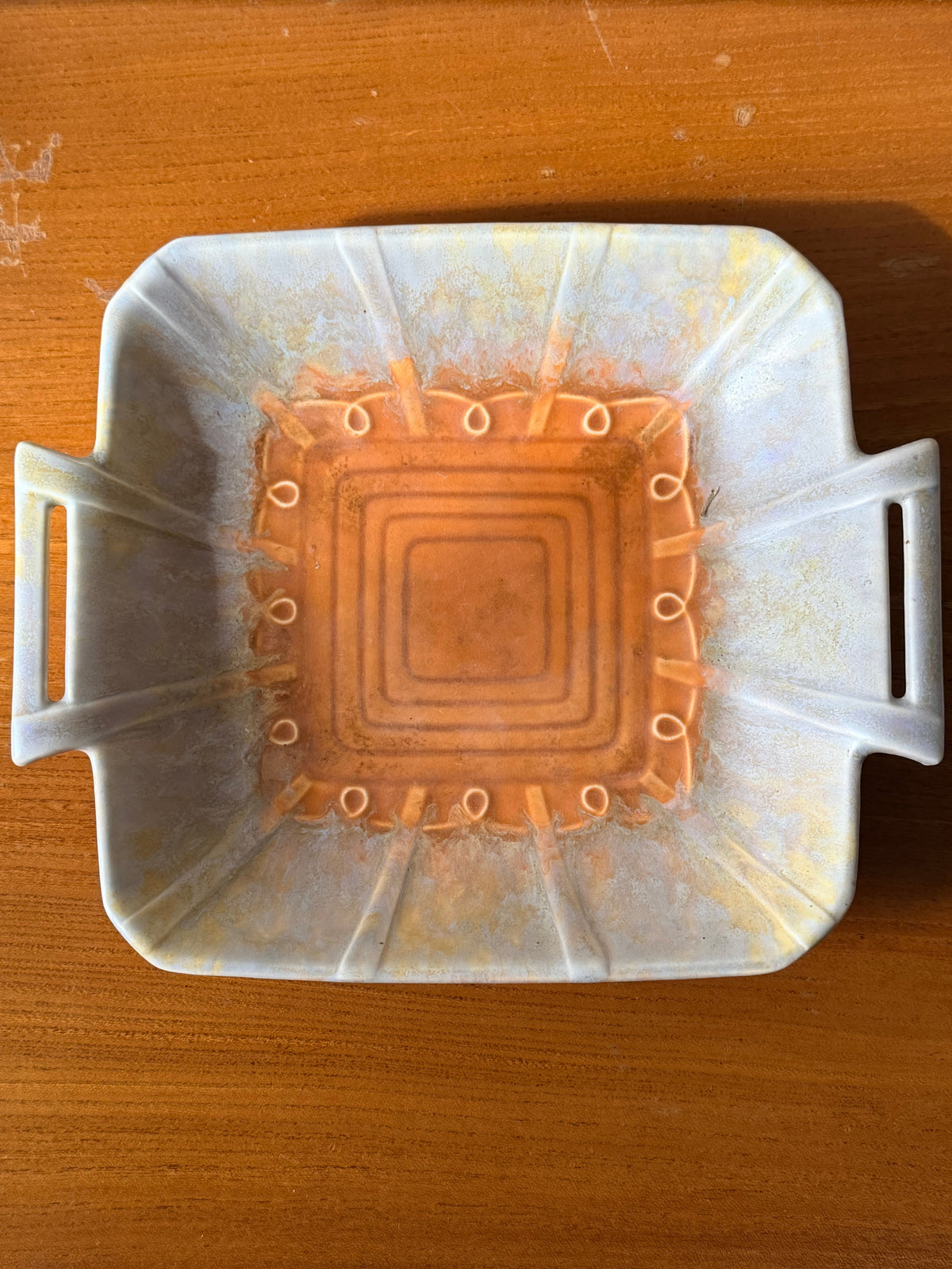 A beswick plate with side handles, featuring a taupe exterior with subtle yellow speckles and a vibrant  burnt orange interior with concentric square patterns and small circular details.
