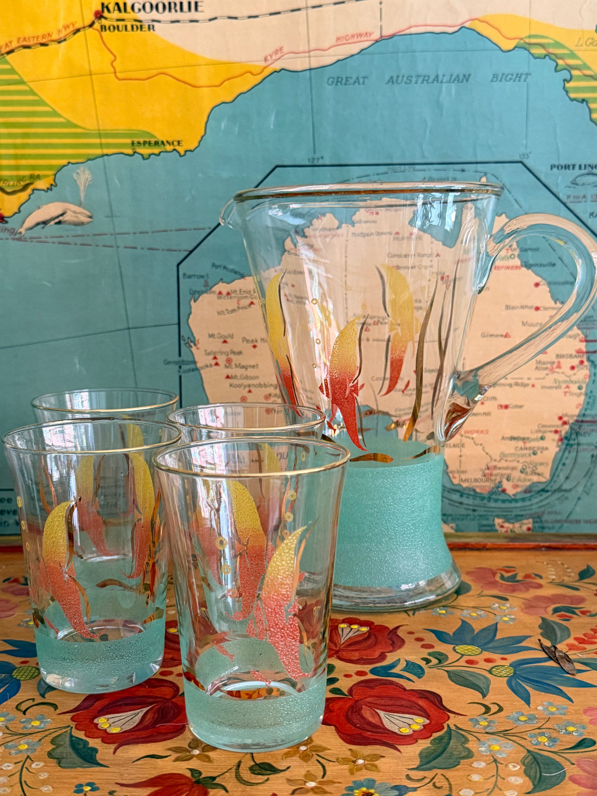 A  retro frosted pitcher with fotumblers, decorated with colorful fish illustrations in yellow, orange, and red and with a sea green frosted band